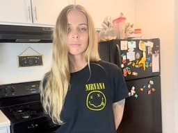 Young woman with long blonde hair stands in a kitchen wearing a Nirvana T-shirt. The kitchen has white cabinets, a black stove, and a fridge with magnets.