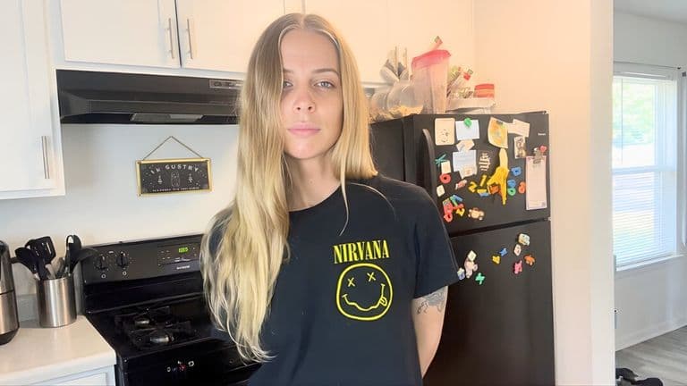 Young woman with long blonde hair stands in a kitchen wearing a Nirvana T-shirt. The kitchen has white cabinets, a black stove, and a fridge with magnets.