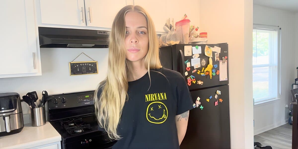 Young woman with long blonde hair stands in a kitchen wearing a Nirvana T-shirt. The kitchen has white cabinets, a black stove, and a fridge with magnets.