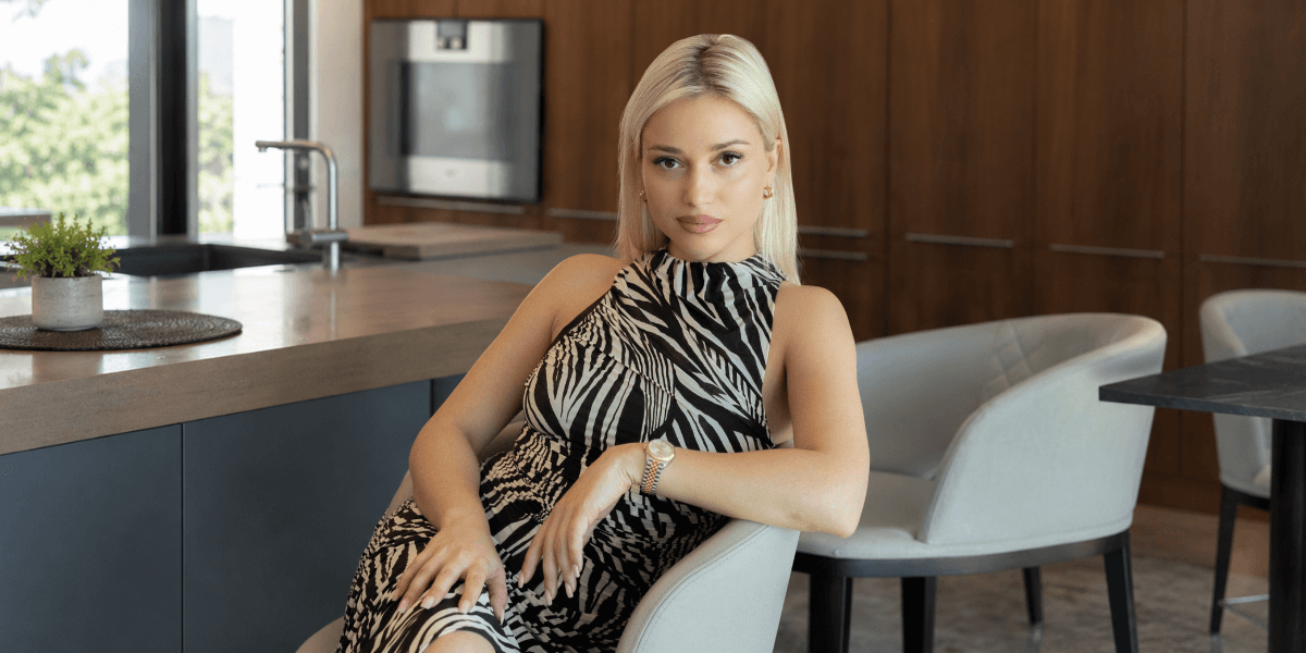 A seated woman with blonde hair poses in a stylish kitchen setting.