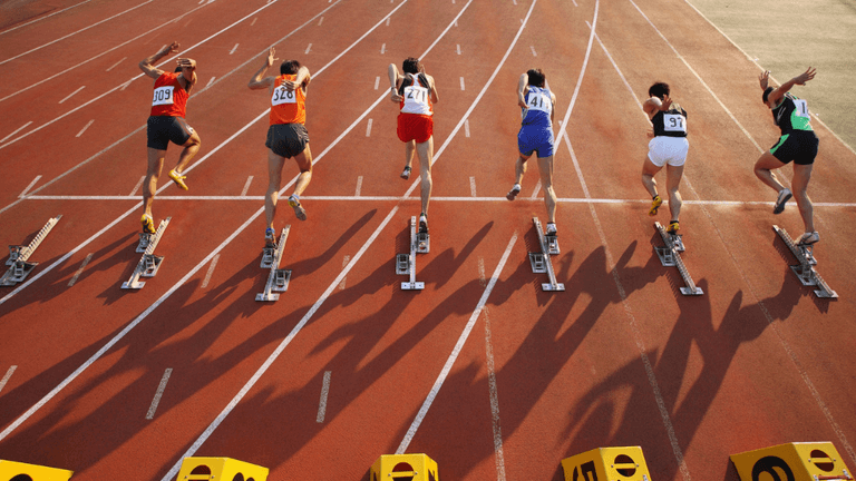 Track and field athletes sprint from starting blocks during a race at an outdoor stadium.