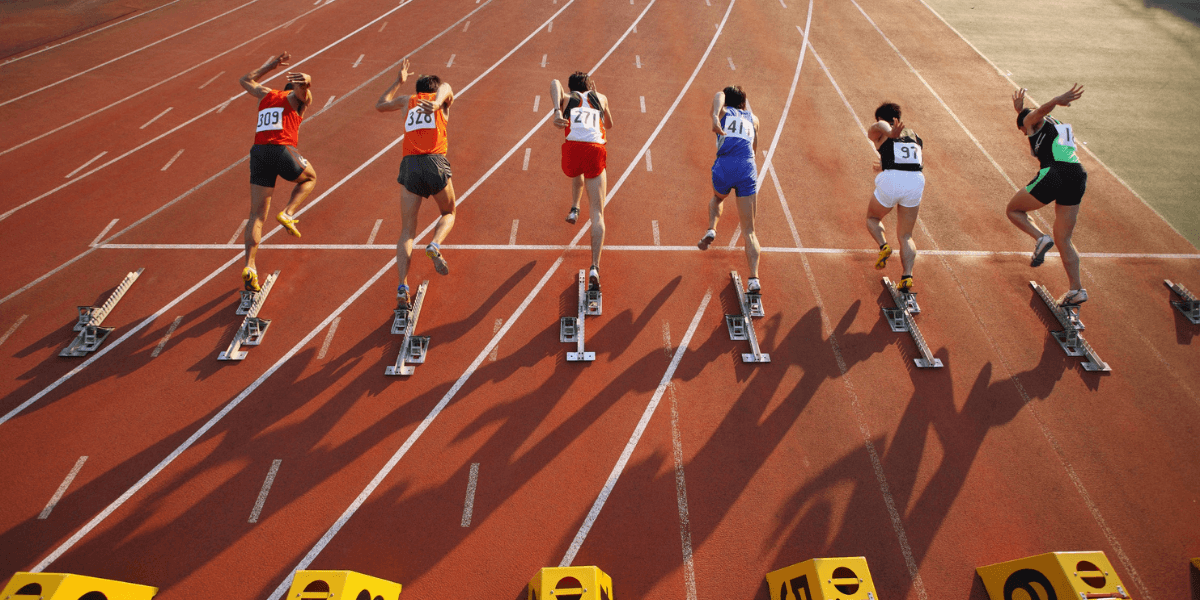Track and field athletes sprint from starting blocks during a race at an outdoor stadium.