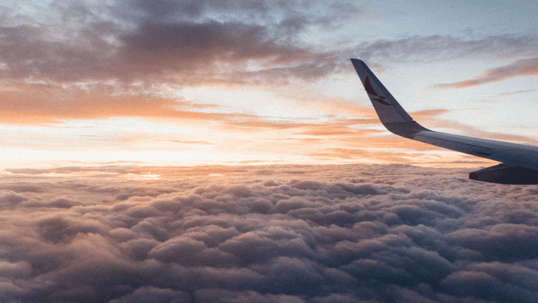 View from an airplane window above the clouds during sunrise with an aircraft wing visible.