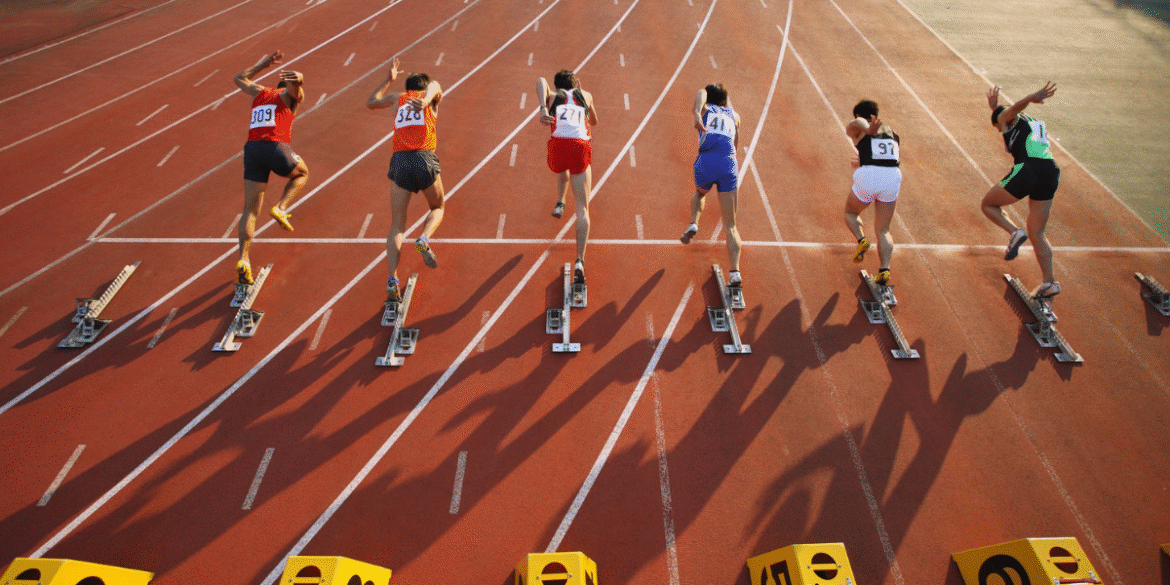 Track and field athletes sprint from starting blocks during a race at an outdoor stadium.