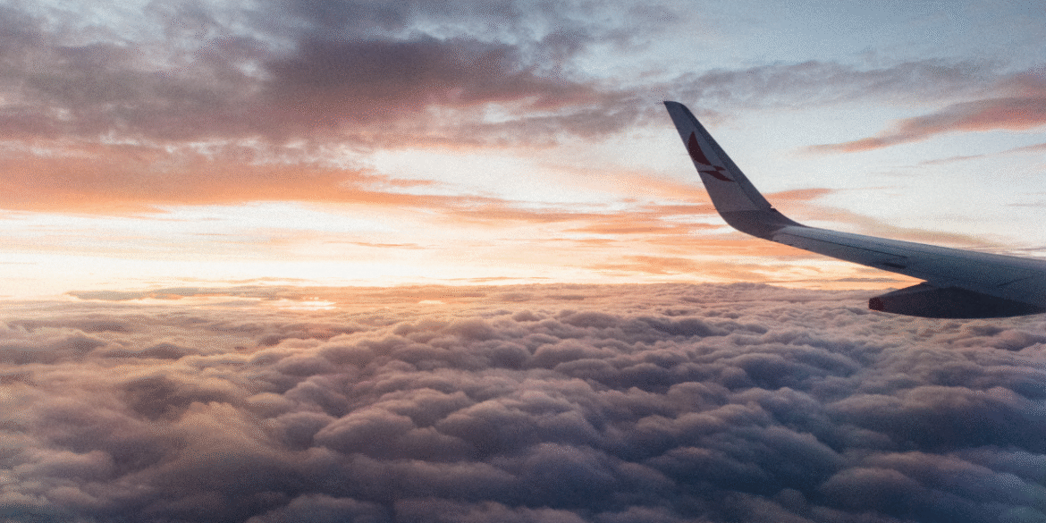 View from an airplane window above the clouds during sunrise with an aircraft wing visible.